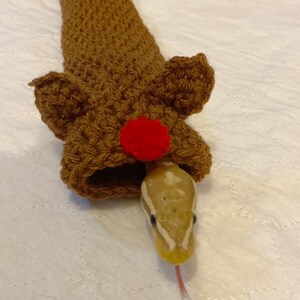 Op de afbeelding: Een bruine, gehaakte rendier-vormige slangschuilplaats met een rode pompon op de neus. Een slang kijkt uit de schuilplaats.