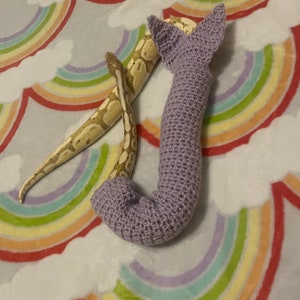 Könnte beinhalten: Ein helllila gehäkeltes Schlangenspielzeug mit einer Schlange, die sich darum windet. Das Schlangenspielzeug liegt auf einer Decke mit Regenbogenmuster.