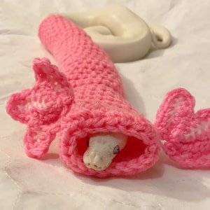 Puede incluir: Una serpiente blanca con ojos rosas está enroscada dentro de una bolsa de dormir de serpiente de crochet rosa con un lazo de crochet rosa en cada extremo.