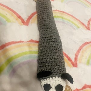 May include: A grey crocheted snake toy with a white crocheted opossum head. The opossum has black eyes and a pink nose.