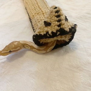 Op de afbeelding: Een beige en zwart gehaakt slangenknuffel met een slangenkop die eruit komt. De slangenknuffel is gemaakt van een zacht, getextureerd garen en heeft een zwarte rand rond de kop. De slang is lichtbruin met donkerdere bruine markeringen.