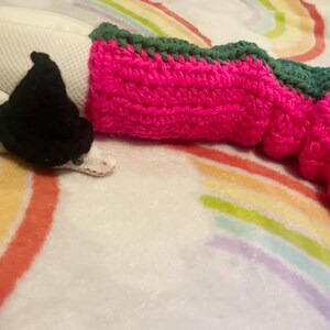 Könnte beinhalten: Eine weiße Schlange mit einem schwarzen gehäkelten Hut und einem rosa-grünen gehäkelten Ärmel. Die Schlange liegt auf einer regenbogenfarbenen Decke.