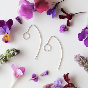 May include: A pair of silver hoop earrings with a simple, curved design. The earrings are photographed against a white background with purple and pink flowers.