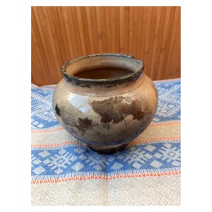 May include: A handmade, round ceramic pot with a wide opening. The pot has a light brown base with darker brown and gray patches. The rim is dark brown. It sits on a blue and white patterned cloth.