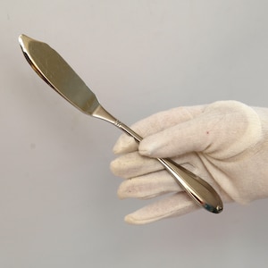 Puede incluir: Un cuchillo de mantequilla plateado con un mango ligeramente curvo se sostiene en un guante de algodón blanco.