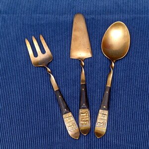 May include: A three-piece set of brass serving utensils including a three-tined fork, a flat cake server, and a spoon. Each piece features a twisted brass neck, a dark brown handle, and a rectangular brass inlay at the base with embossed animal figures.