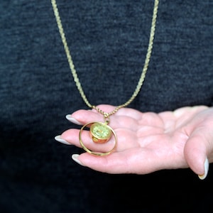 Könnte beinhalten: Eine goldfarbene Halskette mit einem kleinen Uhrenanhänger. Die Uhr hat ein hellgrünes Zifferblatt und ist in einen kreisförmigen Goldrahmen eingefasst. Die Kette ist im zarten Seil-Stil gehalten. Der Hintergrund ist dunkelgrau.