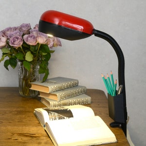 May include: A red and black desk lamp with a flexible neck is positioned over a wooden desk. A vase of purple roses, books, pencils, and an open book are also on the desk. The lamp is turned on, illuminating the desk.