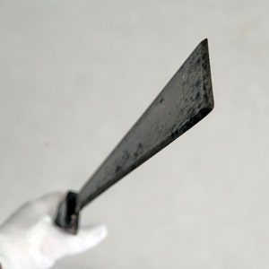 Pode incluir: Um facão escuro e desgastado com uma lâmina retangular e um cabo curto. A lâmina é angulada e parece ser de metal. O facão é segurado por uma mão enluvada.