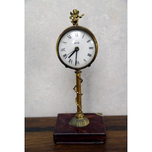 May include: A vintage gold-toned desk clock with a white face and black Roman numerals. The clock has a small cherub figure on top and is mounted on a decorative stand with a marble base. The clock face reads "RITZ".