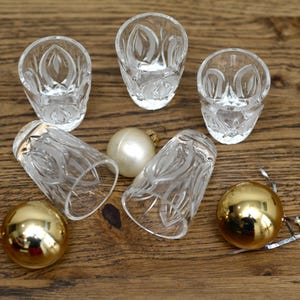 May include: Six clear crystal shot glasses with a leaf design, along with three Christmas ornaments. Two gold ornaments and one white ornament are visible. The shot glasses are arranged on a wooden surface.
