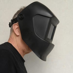 May include: A black welding helmet with a dark viewing window. The helmet has an adjustable head strap and a curved design to protect the face and neck. The helmet is designed for welding and other industrial applications.