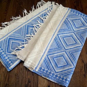 May include: A woven textile featuring a repeating pattern of blue diamonds and geometric shapes on a light beige background. The fabric includes decorative fringe along the edges, with a textured weave that highlights the contrast between the blue and beige.