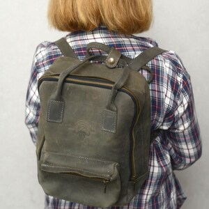 Puede incluir: Una mochila de cuero gris con asa superior y correas ajustables. La mochila tiene un bolsillo frontal con cremallera y un compartimento principal con cierre de cremallera. La bolsa tiene un logotipo en relieve en la parte delantera.