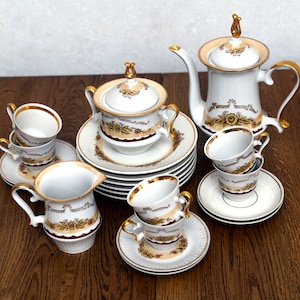 Puede incluir: Un juego de té de porcelana blanca vintage con ribetes dorados y detalles florales. El juego incluye una tetera, azucarero, jarrita de leche, tazas y platillos. Las tazas y platillos están apilados.