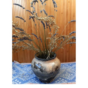 Op de afbeelding: Een ronde, geglazuurde keramieken pot met een gevlekte grijs en beige afwerking, gevuld met een boeket van gedroogde paarse lavendeltakjes en lichtbruine, gestructureerde botanische stengels met fijne bloemtrossen.