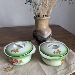May include: Two vintage, light green enamelware bowls with lids, decorated with fruit motifs. The lids have white handles. A brown vase with dried flowers sits in the background on a wooden table.
