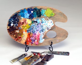 May include: An oval wooden artist palette featuring various dabs of oil paint in shades of blue, green, yellow, red, and white. The palette rests on a black metal stand, accompanied by a metal palette knife and several paint tubes.