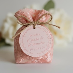 Può includere: Un piccolo sacchetto di pizzo rosa legato con spago, con un'etichetta circolare con le parole "Wedding Carlo & Daniela" e la data 10/05/26. Il sacchetto è pieno e raccolto in alto.