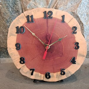 May include: A round wall clock crafted from a natural wood slice featuring a reddish-brown center and a lighter outer ring. The clock face displays black raised numerals from one to twelve, gold-toned hour and minute hands, and a red second hand.