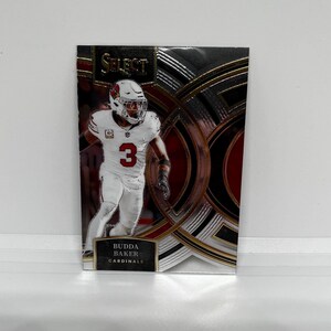 May include: A football trading card featuring a player in a white uniform with the number 3 on the front. The card has a silver, gold, and red background with the text "SELECT" and "BUDDA BAKER CARDINALS".