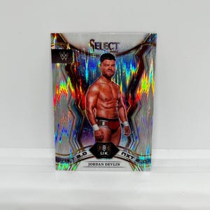 May include: A silver and gold WWE Select 2022 trading card featuring a wrestler with a black and red wrestling outfit. The card has a holographic background with a rainbow effect. The wrestler's name is Jordan Devlin and the card is labeled NXT 2.0.