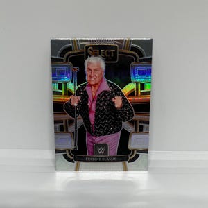 May include: A silver, gold, and black trading card featuring a portrait of Freddie Blassie, a professional wrestler. The card has a holographic background and the text "SELECT" and "FREDDIE BLASSIE" on it.