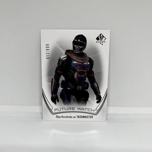 May include: A white trading card with a black and white image of a character wearing a black helmet and a black and red suit. The card has the text "FUTURE WATCH" and "Olga Kurylenko as TASKMASTER" on it. The card also has the number "612/999" on it.