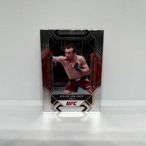 May include: A silver and black trading card featuring a photo of Askar Askarov, a UFC flyweight fighter, in a fighting stance. The card has a red and gold border and the text "UFC" is printed on the card.