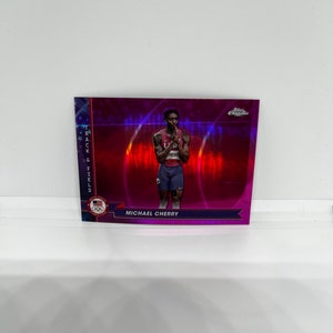 Puede incluir: Una tarjeta coleccionable deportiva con Michael Cherry en uniforme rojo y azul. La tarjeta tiene una superficie brillante y reflectante con las palabras "Track & Field" y "Chrome" visibles. La tarjeta también presenta el logo olímpico de EE. UU.