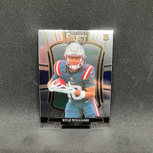 May include: A football trading card featuring Kyle Williams of the Patriots. The card has a silver and blue design with the text "SELECT" at the top. Williams is wearing a helmet and holding a football. The card also has the text "RC" and "2023".
