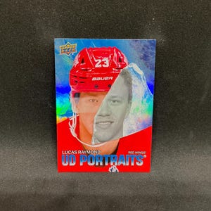 May include: A hockey trading card featuring Lucas Raymond of the Red Wings. The card has a portrait of the player, with a red helmet and the number 23. The card is labeled "UD Portraits".