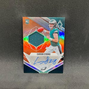 May include: A Carson Strong football trading card featuring a holographic orange and teal background, a rookie card logo, a green fabric jersey swatch, and a blue ink autograph. The player is shown in a Philadelphia Eagles uniform holding a football.