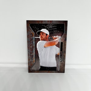 Puede incluir: Una tarjeta de intercambio con un golfista con una camisa blanca y una gorra negra, balanceando un palo de golf. La tarjeta tiene un fondo metálico con las palabras "The Metalized" y "Matthew Wolff" impresas en ella.