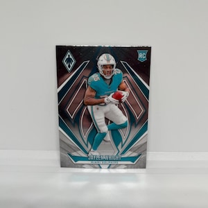 Puede incluir: Una tarjeta de coleccionista de fútbol con Jaylen Wright de los Miami Dolphins. La tarjeta tiene un diseño de mosaico con acentos turquesa, blancos y negros. Wright está representado con su uniforme, sosteniendo un balón de fútbol. La tarjeta incluye el logo RC.
