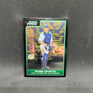 Puede incluir: Una tarjeta de béisbol de Russ Martin de los Dodgers de Los Ángeles. La tarjeta presenta una imagen en color del jugador con su uniforme de receptor. La tarjeta tiene un borde verde y las palabras "Bowman Chrome" en la parte superior.