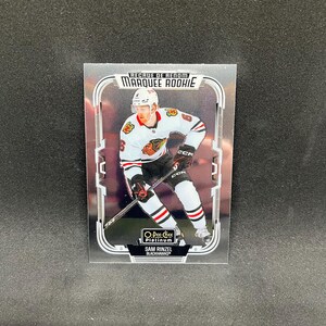 Może przedstawiać: Prostokątna karta sportowa przedstawiająca hokeistę w białym stroju Chicago Blackhawks z czarnymi i czerwonymi akcentami. Na karcie widnieje napis Marquee Rookie, O-Pee-Chee Platinum oraz imię i nazwisko zawodnika Sam Rinzel na dole.