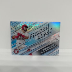 May include: Baseball trading card featuring Kyle Schwarber of the Philadelphia Phillies. The card has a holographic design with the words "Frozen Ropes" in a stylized font. The player is shown in action, wearing a red helmet and batting gloves.