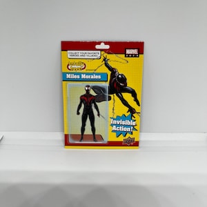 K&ouml;nnte beinhalten: Eine schwarze und rote Miles Morales Actionfigur vor gelbem Hintergrund. Die Figur ist in einer Web-Schwing-Actionpose dargestellt. Die Verpackung enth&auml;lt den Text "Totally Toys! Presents Miles Morales" und "Invisible Action!"