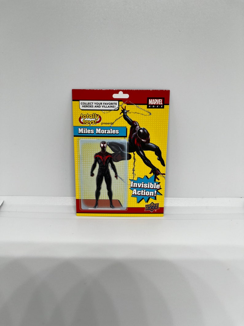 K&ouml;nnte beinhalten: Eine schwarze und rote Miles Morales Actionfigur vor gelbem Hintergrund. Die Figur ist in einer Web-Schwing-Actionpose dargestellt. Die Verpackung enth&auml;lt den Text "Totally Toys! Presents Miles Morales" und "Invisible Action!"