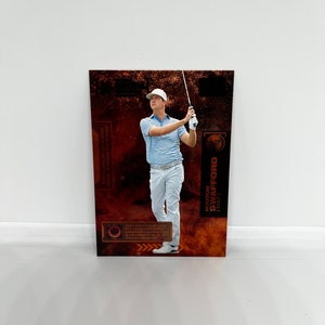 Puede incluir: Una tarjeta de intercambio con un golfista con una camisa blanca y pantalones blancos, sosteniendo un palo de golf. La tarjeta tiene un fondo marrón y naranja con el texto "HUDSON SWAFFORD | GOLF" en blanco.