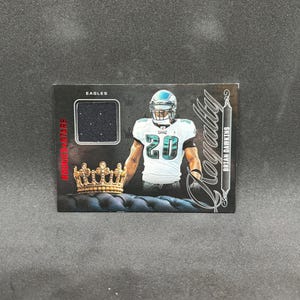May include: A rectangular sports trading card featuring Brian Dawkins in a white Philadelphia Eagles jersey with the number 20. The card includes a dark fabric swatch, a gold crown graphic, and the text Eagles, Royalty, and Rookie Stars.