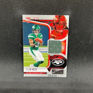 May include: A New York Jets football trading card featuring Elijah Moore, a wide receiver. The card has a green, white, and red color scheme, with the Jets logo and the text "PLAYBOOK". The card also includes the text "ROOKIE CARD".