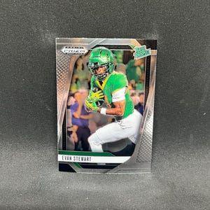 Puede incluir: Una tarjeta de fútbol coleccionable Panini Prizm con Evan Stewart con uniforme verde y blanco. La tarjeta tiene un borde plateado y las palabras "Rated Prospect" son visibles. El jugador sostiene un balón de fútbol.