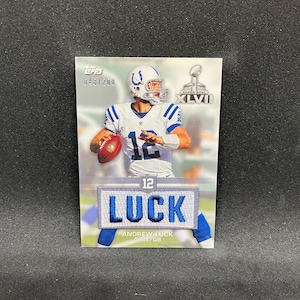 May include: A Topps football card featuring Andrew Luck of the Indianapolis Colts. The card shows Luck in his Colts uniform, with the Super Bowl XLVI logo. The card also has a patch with the name "LUCK" and the number 12.