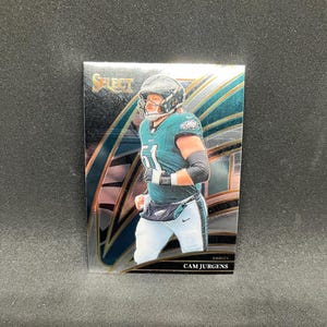 May include: A football trading card featuring Cam Jurgens of the Philadelphia Eagles. The card has a holographic design with teal and silver accents. Jurgens is wearing a teal jersey with the number 51 and white trousers.
