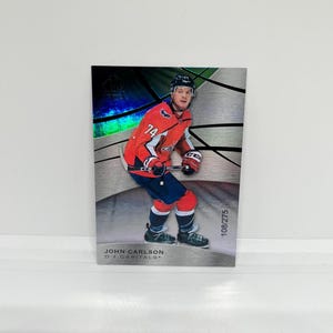 May include: A hockey trading card featuring John Carlson, a defenceman for the Washington Capitals. The card has a silver background with a holographic effect and the player's number 74 is visible on his jersey.