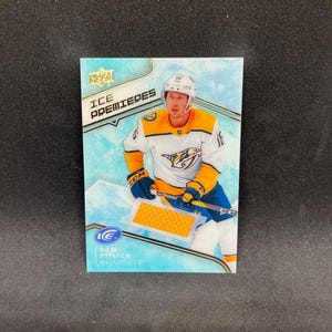 May include: A sports trading card featuring hockey player Rem Pitlick in a white and yellow Nashville Predators jersey. The card includes an embedded yellow fabric swatch, the text ICE PREMIERES, and the player's name and team at the bottom.