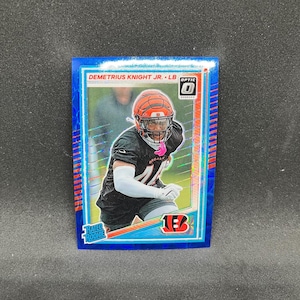 Pode incluir: Um cartão de negociação de futebol Optic azul com Demetrius Knight Jr. - LB. O cartão mostra um jogador em uniforme preto e laranja, usando um capacete. O cartão tem um logotipo Rated Rookie e o logotipo da equipe Bengals.