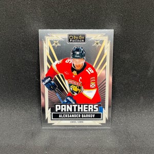 Może przedstawiać: Karta hokejowa O-Pee-Chee Platinum przedstawiająca Aleksandera Barkova z Florida Panthers. Karta ukazuje Barkova w czerwonej koszulce z numerem 16, trzymającego kij hokejowy. Tekst na karcie brzmi PANTHERS oraz ALEKSANDER BARKOV.